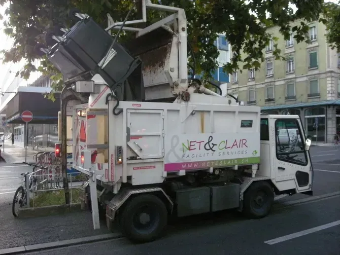 Camion utilitaire blanc avec nacelle élévatrice levée, stationné dans une rue à côté de bâtiments et de vélos.