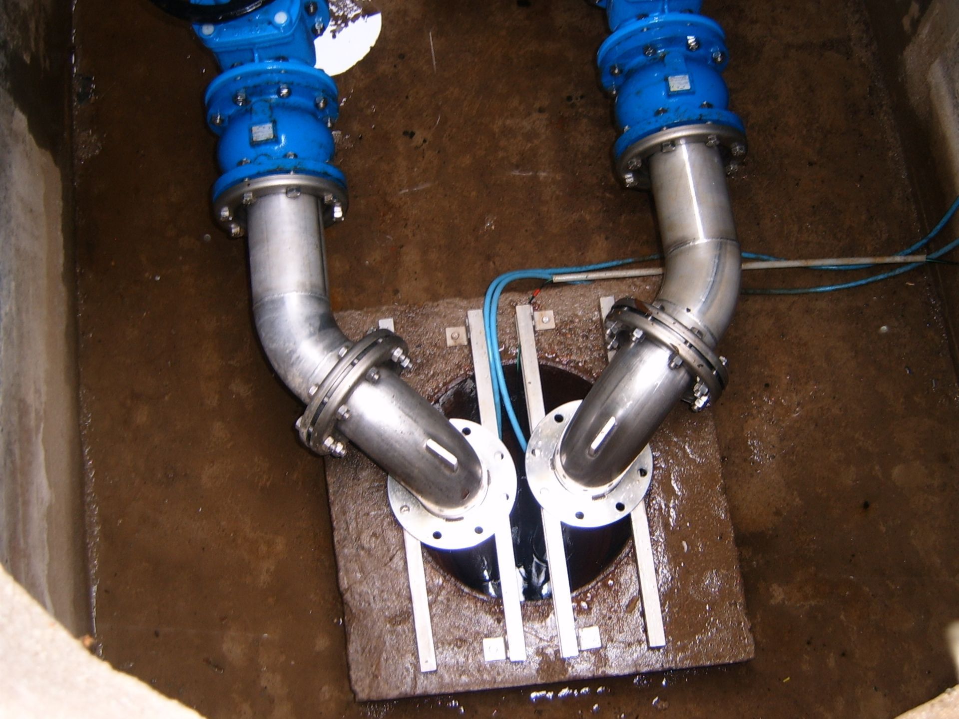 Deux tuyaux à embouts bleus et courbes argentées rejoignent un appareil central noir dans un décor de béton brun humide.
