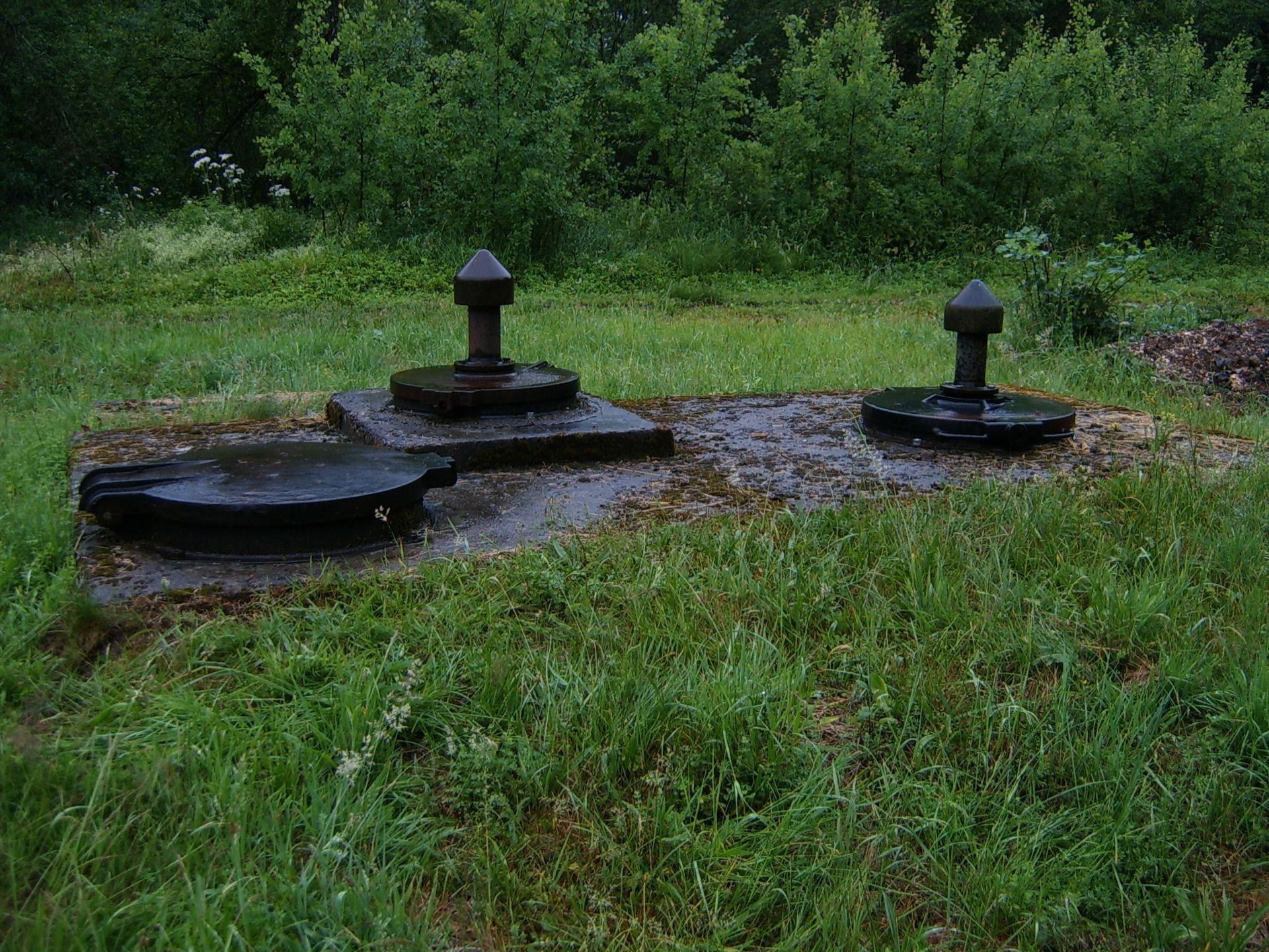 Trois couvercles de soupapes métalliques dans une zone herbeuse, deux avec des boutons en relief.