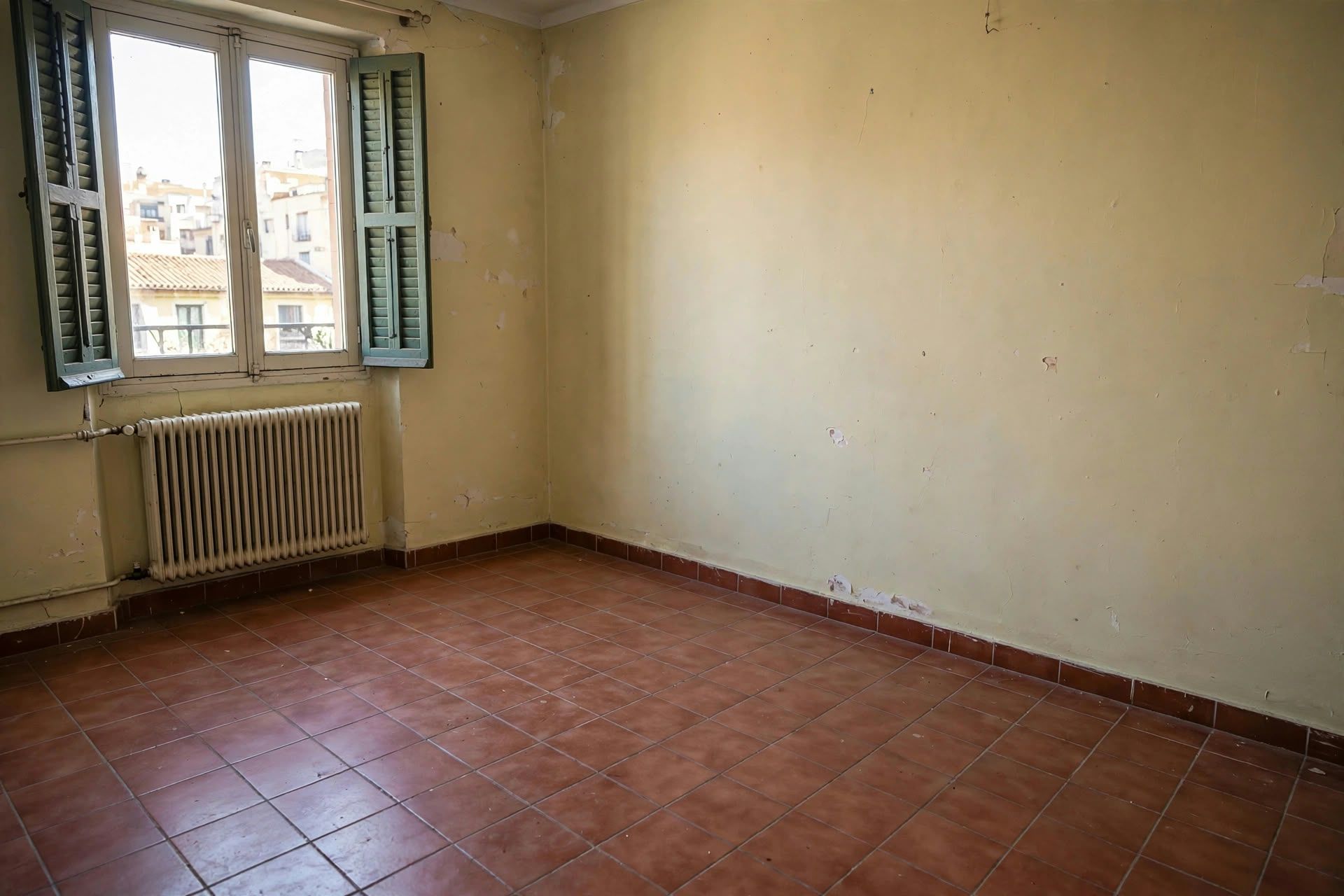 Habitación vacía con suelo de baldosas rojas, paredes beige y una ventana con contraventanas verdes y un radiador debajo
