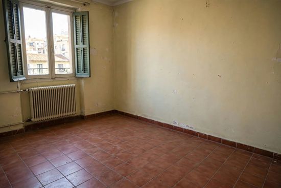 Habitación vacía de color beige, con suelo de baldosas rojas, una ventana con contraventanas verdes y un radiador debajo