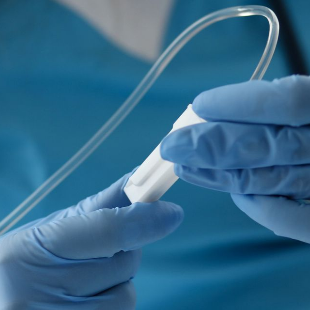 Des mains gantées de gants en nitrile bleus tiennent un petit dispositif médical blanc relié à un tube en plastique transparent.