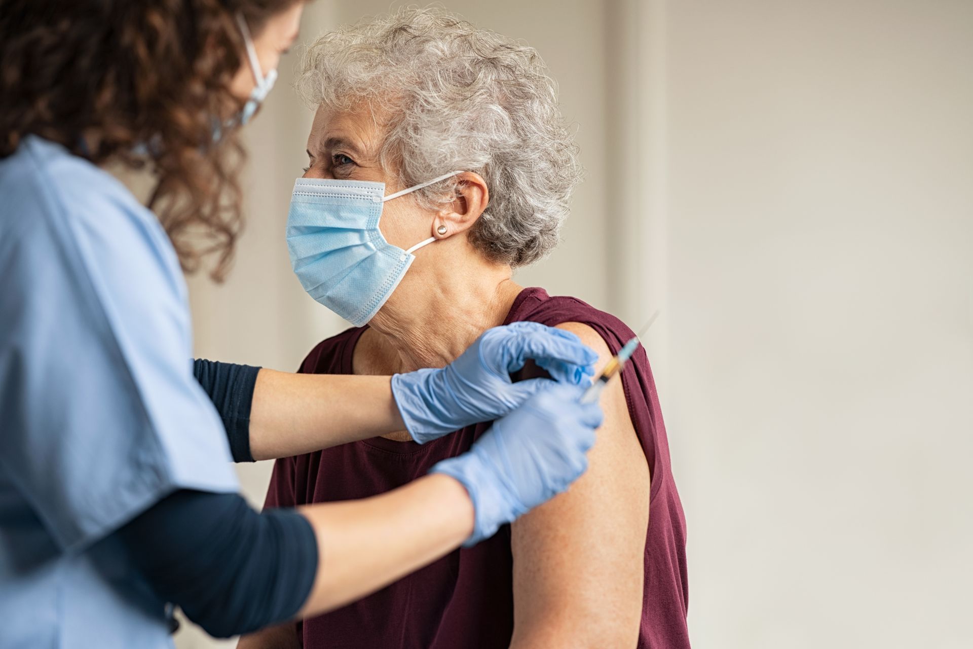 Une infirmière vêtue d'une blouse bleue et d'un masque administre une injection de vaccin dans l'épaule d'une personne.