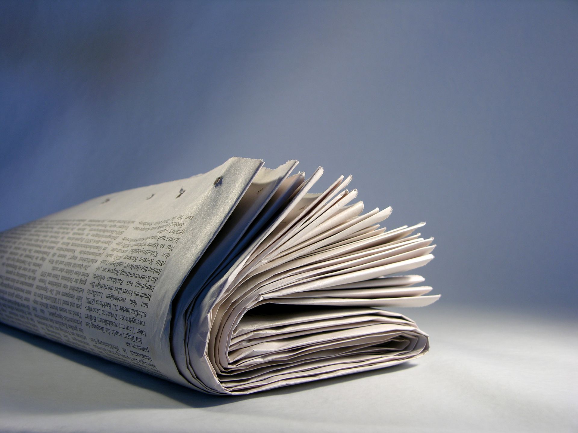 Gefaltete Zeitung auf weißem Untergrund, bedruckte Seiten sichtbar, vor blauem Hintergrund.