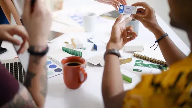 Personas examinando una tarjeta de presentación en una mesa, rodeadas de suministros de oficina, café y elementos de diseño, intercambiando ideas en un entorno bien iluminado.