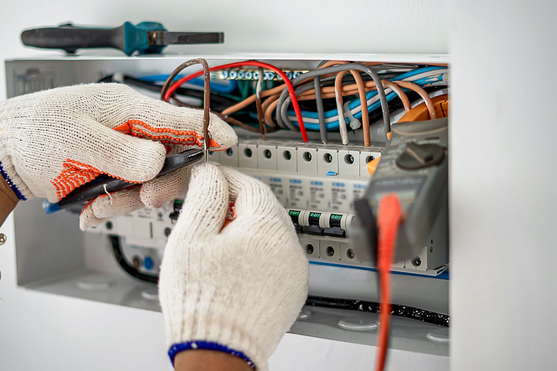 Des mains gantées câblent un tableau de disjoncteurs avec des câbles orange et bleus dans une armoire électrique