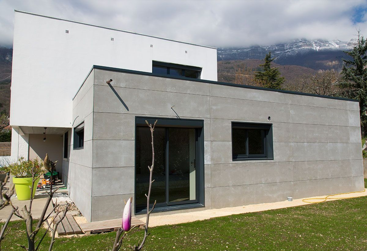 Maison moderne blanche et grise avec une petite pelouse et une vue sur les montagnes en arrière-plan.