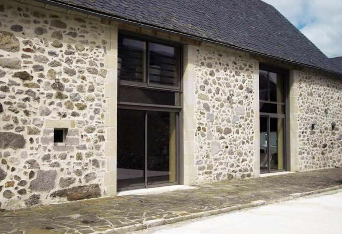 Façade en pierre d'un bâtiment avec des fenêtres à encadrement sombre et une porte en retrait sous un toit en pente.