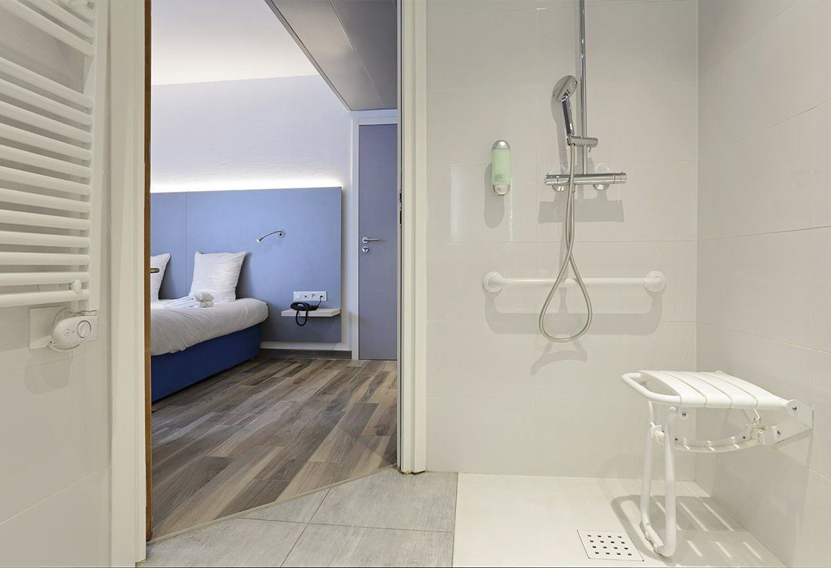 Petite salle de bains blanche avec douche, à côté d'une porte ouverte donnant sur une chambre bleue avec lit et oreillers.