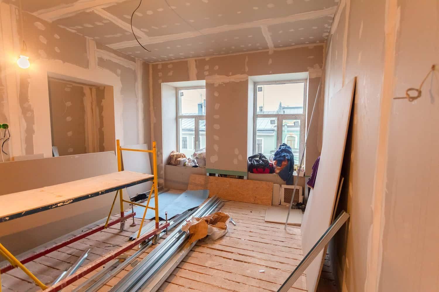 Pièce rénovée en cours de travaux avec cloisons sèches, échelles, outils et plancher en bois apparent près de deux fenêtres