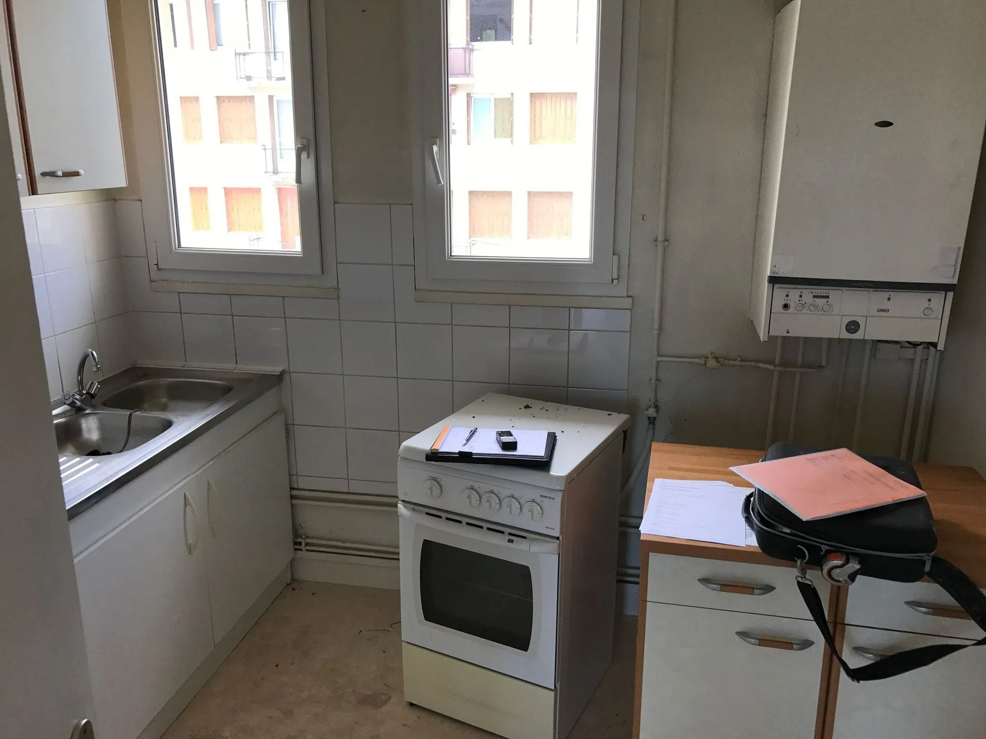 Petite cuisine avec cuisinière, évier, placards et table avec des papiers sous deux fenêtres.