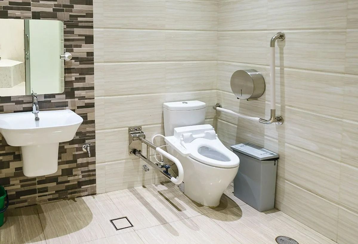 Toilettes accessibles avec lavabo mural, barres d'appui et poubelle, sol et murs carrelés de beige