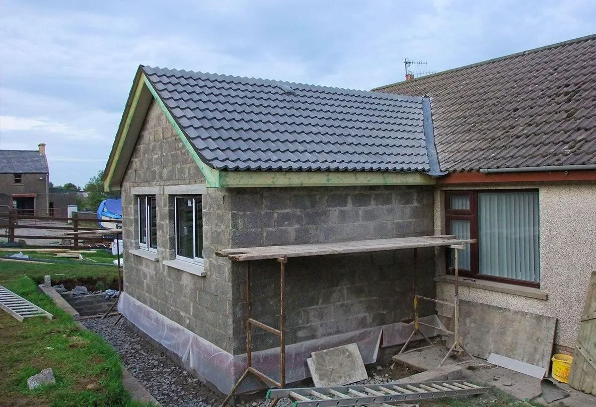 Petite dépendance inachevée au toit gris, jouxtant une maison et entourée d'une cour en désordre.