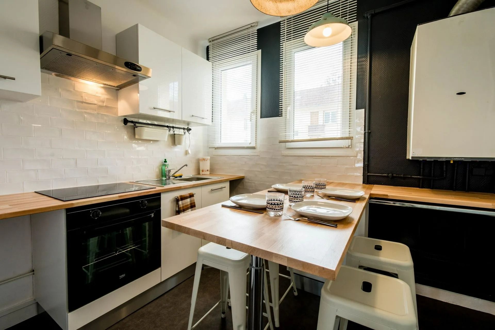 Cuisine moderne avec placards blancs, table en bois dressée pour les repas et deux fenêtres laissant entrer la lumière naturelle.