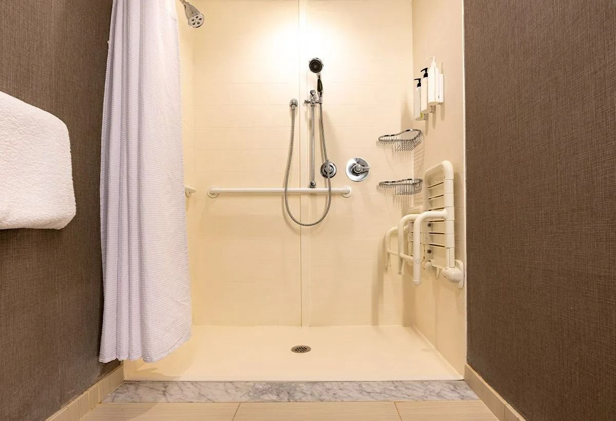 Douche accessible avec barres d'appui, pommeau de douche à main, banc intégré et murs couleur crème.