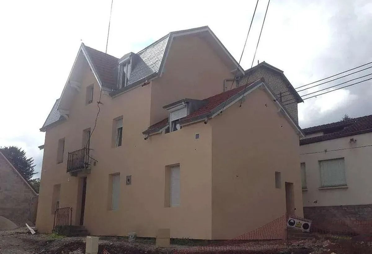 Maison beige à deux étages avec un toit pentu et des lucarnes sous un ciel nuageux