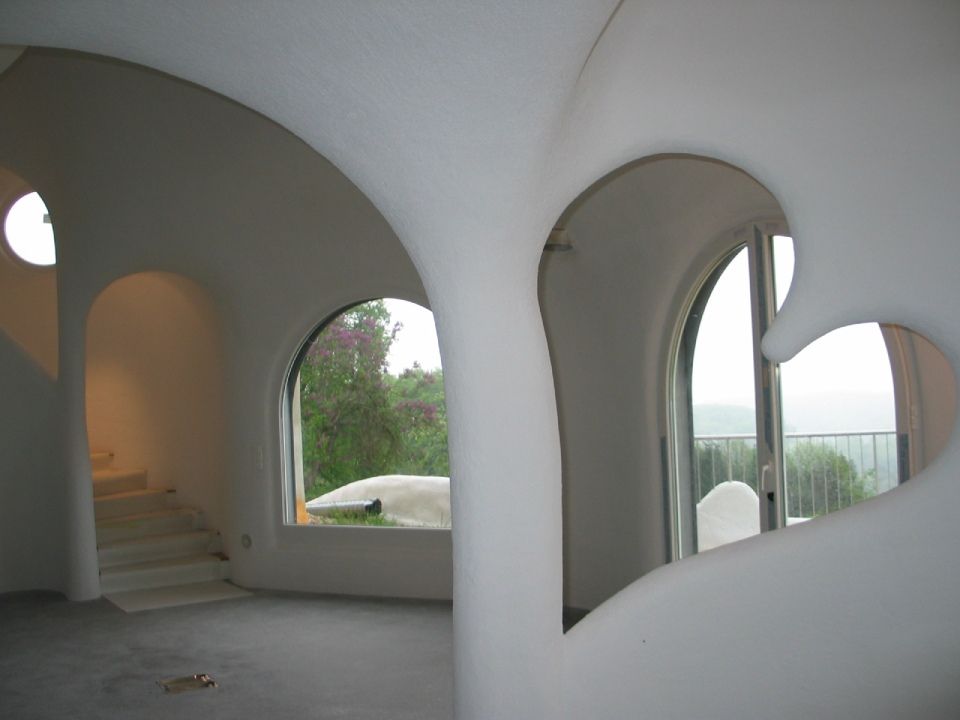Innenansicht eines weissen, organisch geformten Hauses mit Bogenfenstern, Treppe und herzförmiger Öffnung.