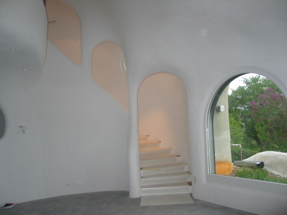 Innenansicht eines weissen Gebäudes mit geschwungenen Wänden, Treppenhaus, Bogentüren und einem grossen Fenster mit Blick ins Freie.