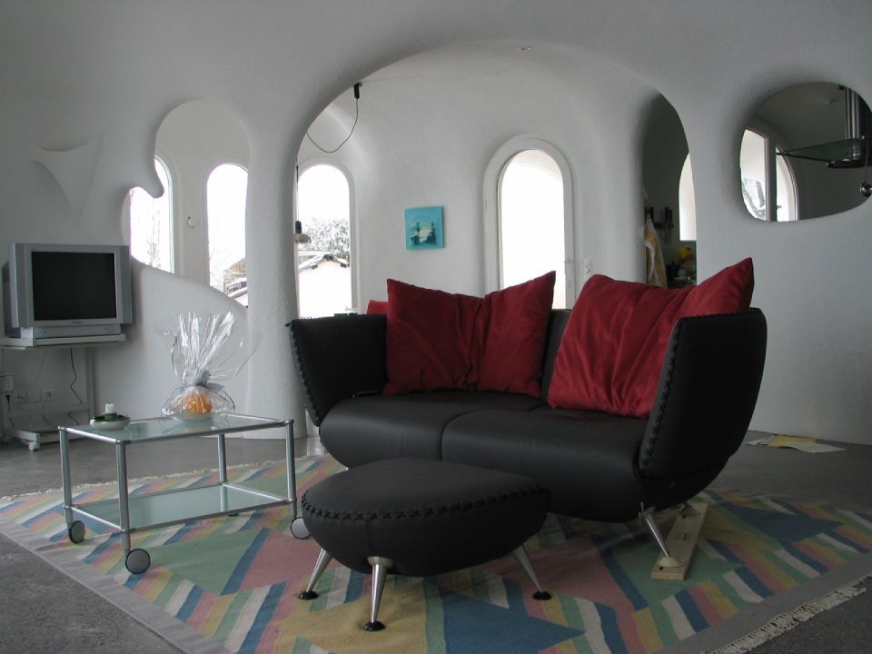 Wohnzimmer mit schwarzem Sofa, roten Kissen, Hocker und buntem Teppich. Weisse, abgerundete Wände.