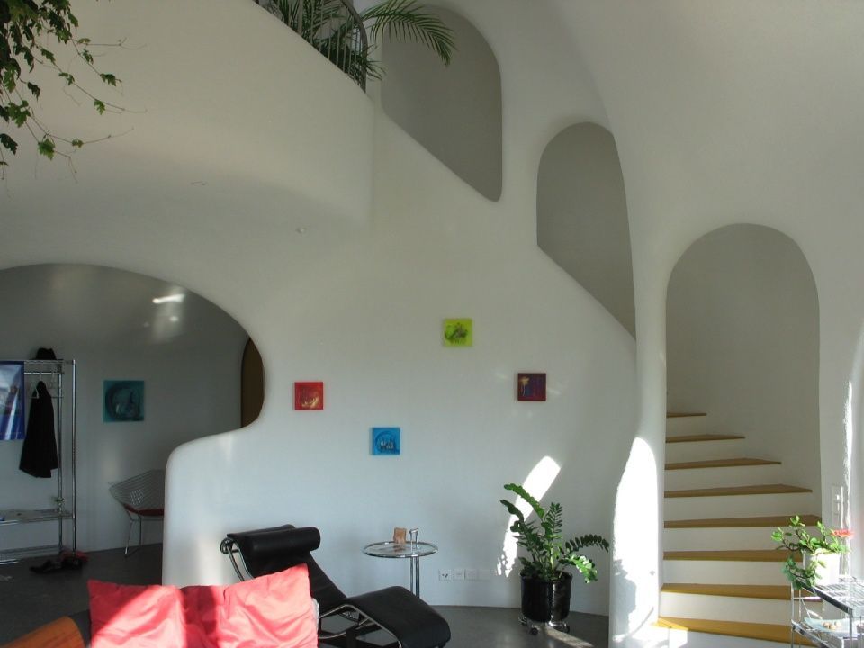 Weisser Innenraum mit geschwungenen Wänden, Treppe und farbenfrohen quadratischen Kunstwerken. Sonnenlicht strömt durch die Torbögen und beleuchtet die Pflanzen.