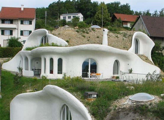 Weisses, organisch geformtes Haus am Hang mit runden Fenstern und einem grasbewachsenen Dach.