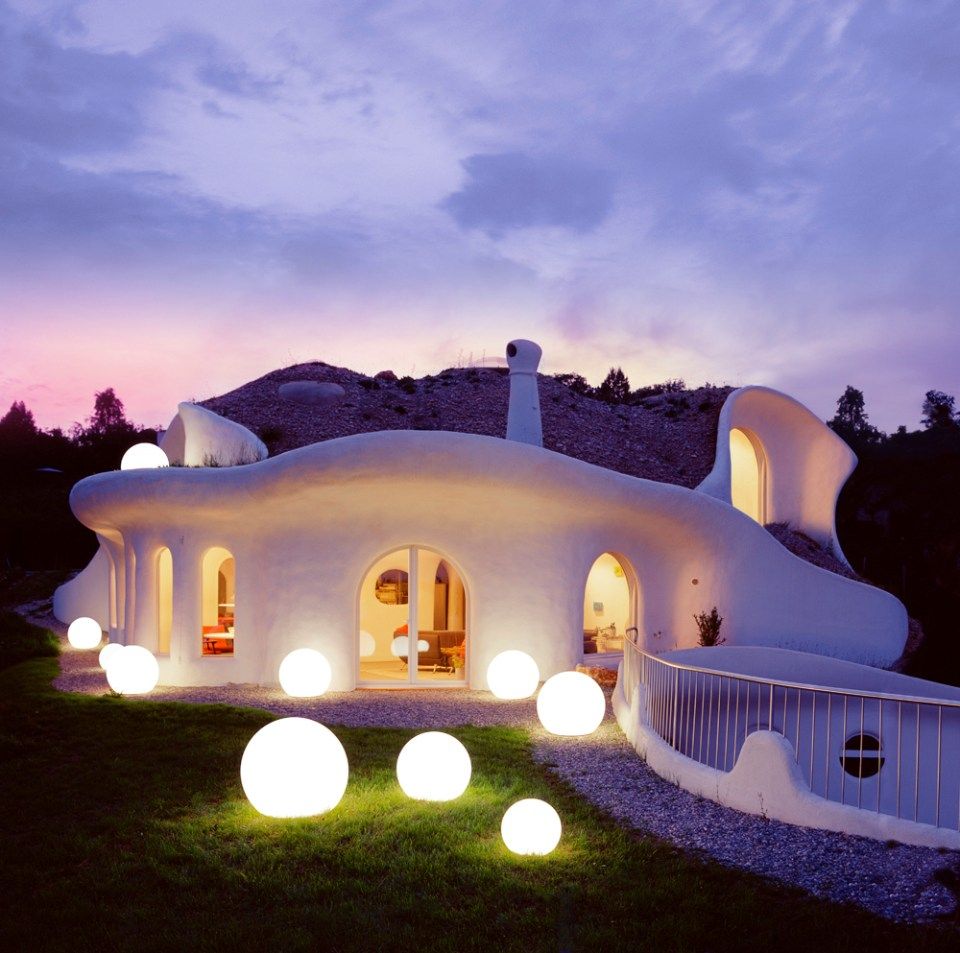Ein weisses, skurriles, in die Erde gebautes Haus mit leuchtenden Kugeln im Garten vor einem dämmernden Himmel.