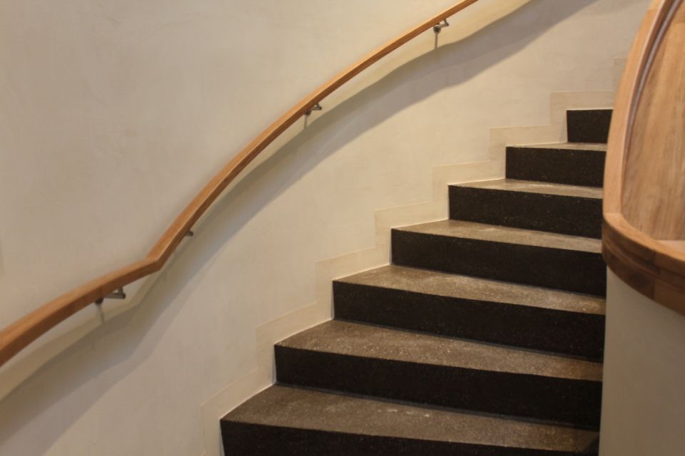 Treppe mit hölzernem Handlauf, der sich an einer weissen Wand entlang nach oben windet. Die Stufen sind dunkelgrau.