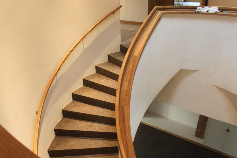 Geschwungene Treppe mit braunen Teppichstufen, hölzernem Handlauf und weissen Wänden.