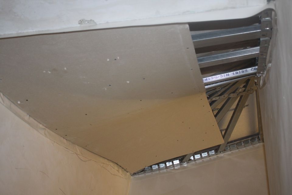 Die Gipskartondecke wurde teilweise entfernt und gibt den Blick auf ein Metallgerüst und einen unfertigen Raum frei. Beige Gipskartonwände, Metallträger und helle Wände.