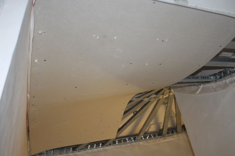 Nahaufnahme einer beschädigten Decke mit sichtbaren Gipskartonplatten und Metallrahmen. Beige- und Weisstöne dominieren.