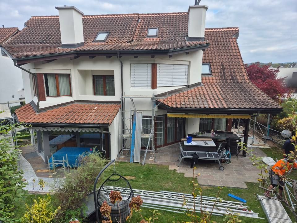 Ein zweistöckiges Haus mit rotem Ziegeldach, Terrasse und Gerüst; im Aussenbereich sind Arbeiten im Gange.