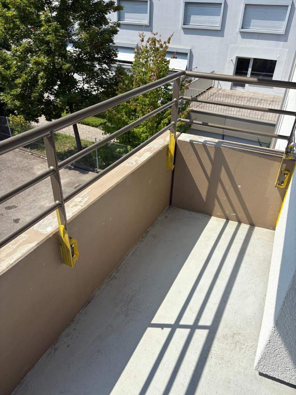 Balkon mit Betonboden, hellbraunen Wänden und Metallgeländer. Helles Sonnenlicht wirft Schatten.