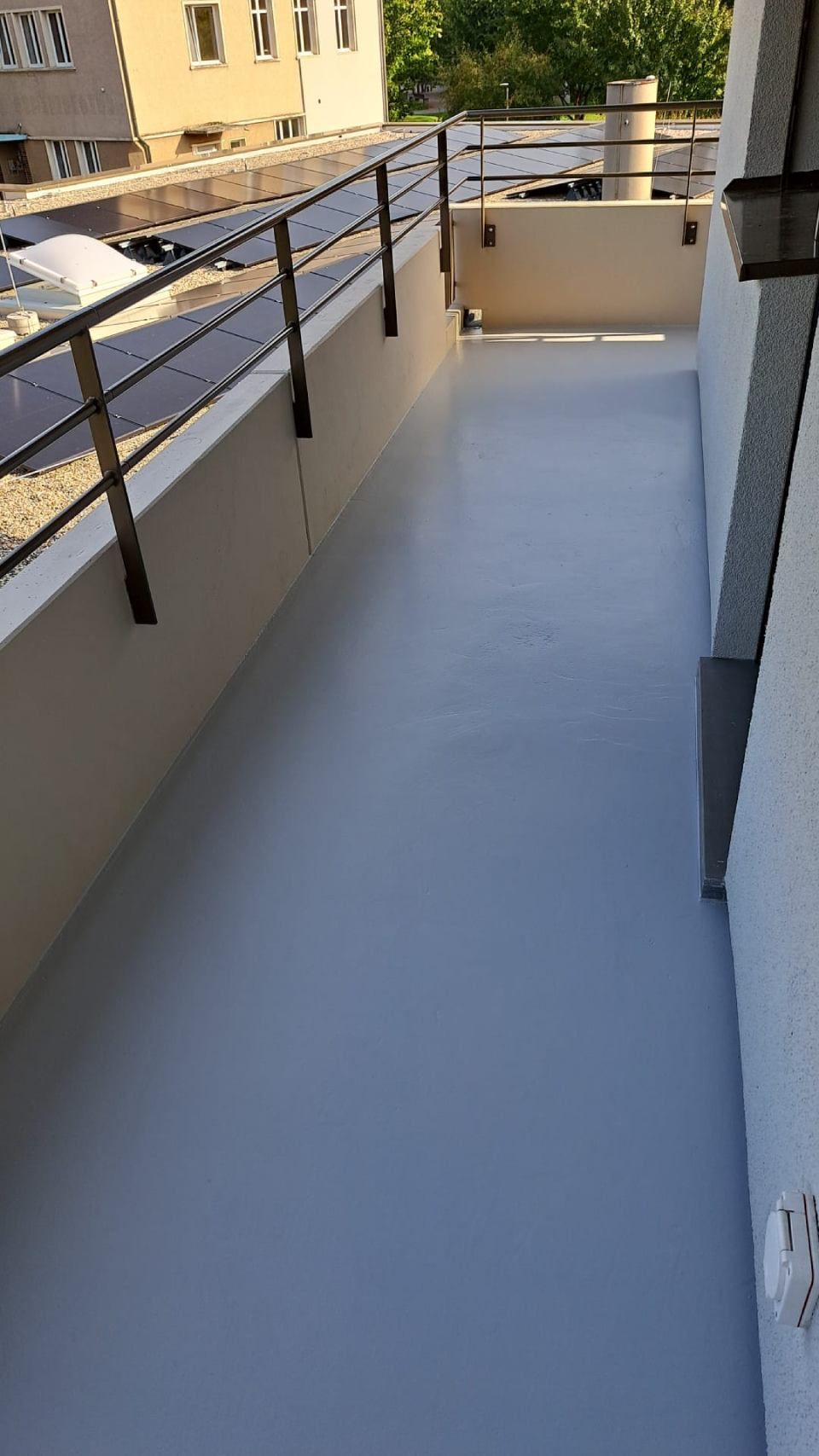 Schmaler Balkon mit grauer, wasserdichter Beschichtung, schwarzem Geländer und Blick auf das Gebäude.