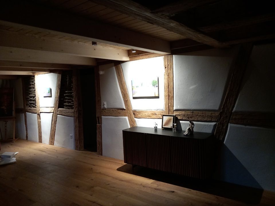 Der Innenraum besticht durch freiliegende Holzbalken, weisse Wände und einen Holzschrank. Sonnenlicht strömt durch die Fenster.