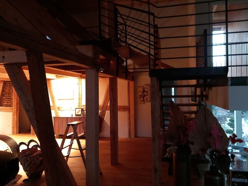 Innenansicht eines zweistöckigen Hauses mit freiliegenden Holzbalken, einer Metalltreppe und einem Fenster, das für natürliches Licht sorgt.