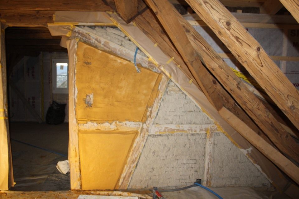 Innenansicht eines Dachbodens mit freiliegenden Holzbalken und teilweise verputzten Wänden in Gelb- und Weisstönen.