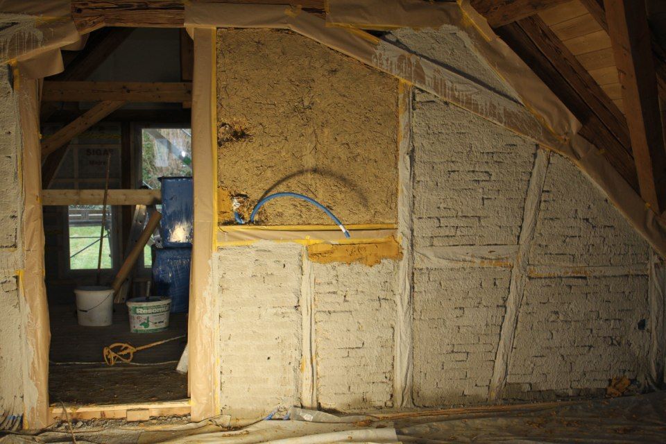 Innenansicht eines im Bau befindlichen Gebäudes. Wand mit strukturiertem Lehmputz und sichtbarem Holzfachwerk; Türöffnung führt ins Freie.