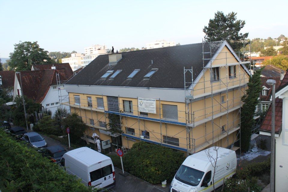 Ein Gebäude wird renoviert; es ist ein Gerüst angebracht, hat gelbe Dämmung und ein dunkles Dach. Mehrere Fahrzeuge und Bäume sind geparkt.