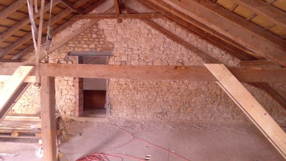 Innenansicht eines Dachbodens mit freiliegenden Holzbalken und Steinwänden, einer Türöffnung und Baumaterialien.