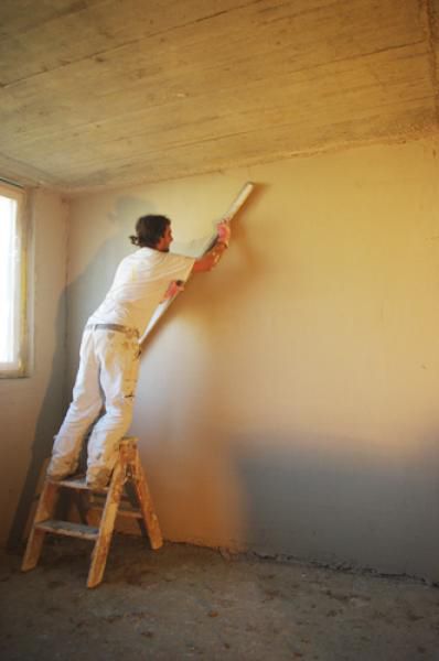 Ein Arbeiter in weissem Overall auf einer Leiter glättet mit einer langen Kelle den Putz an einer Wand. Innenaufnahme.