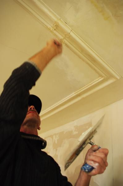 Eine Person auf einer Trittleiter benutzt eine Kelle, um Gips an eine Decke in der Nähe von Zierleisten aufzutragen.