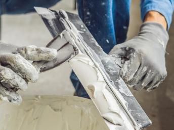 Hände in grauen Handschuhen benutzen eine Kelle, um Fugenmasse aus einem Eimer aufzutragen.