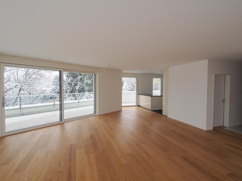 Leeres Zimmer mit Holzboden, grosser Glasschiebetür und Balkon mit Blick auf eine verschneite Landschaft.