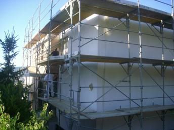 Ein Gerüst umgibt die weisse Fassade eines Gebäudes. Zwei Personen arbeiten an der Seite.