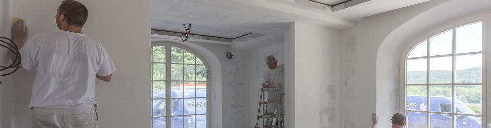 Maler sind bei der Renovierung eines Zimmers beschäftigt und tragen weisse Farbe auf Wände und Decke auf.