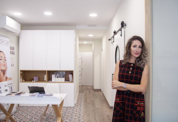 Mujer en una clínica, con los brazos cruzados, de pie junto a una puerta. Armarios blancos, escritorio y colores neutros.