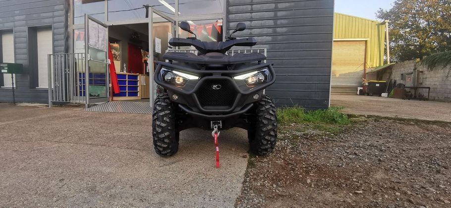 Un quad noir est stationné sur du gravier devant un bâtiment, avec une sangle de remorquage rouge qui pend à l'avant.