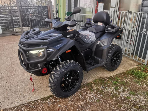 Un quad noir, équipé de pneus à crampons et d'une sangle de remorquage rouge à l'avant, est stationné sur du gravier à côté d'une clôture métallique.