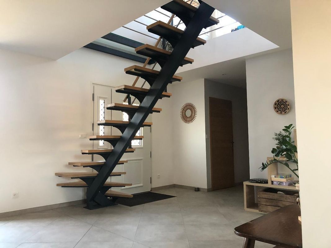 Salon moderne avec escalier flottant noir, puits de lumière, sol carrelé et petite plante près du mur
