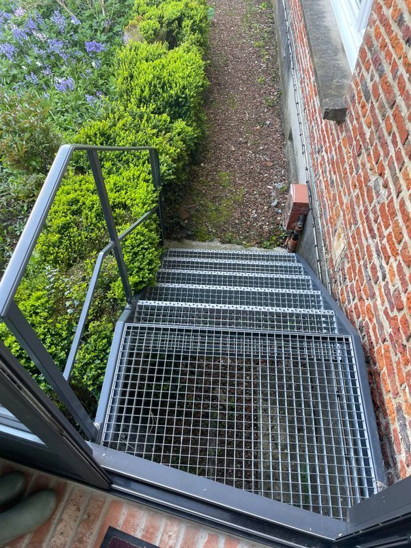 Escalier métallique descendant le long d'un mur de briques et de buissons verts, vu d'en haut.